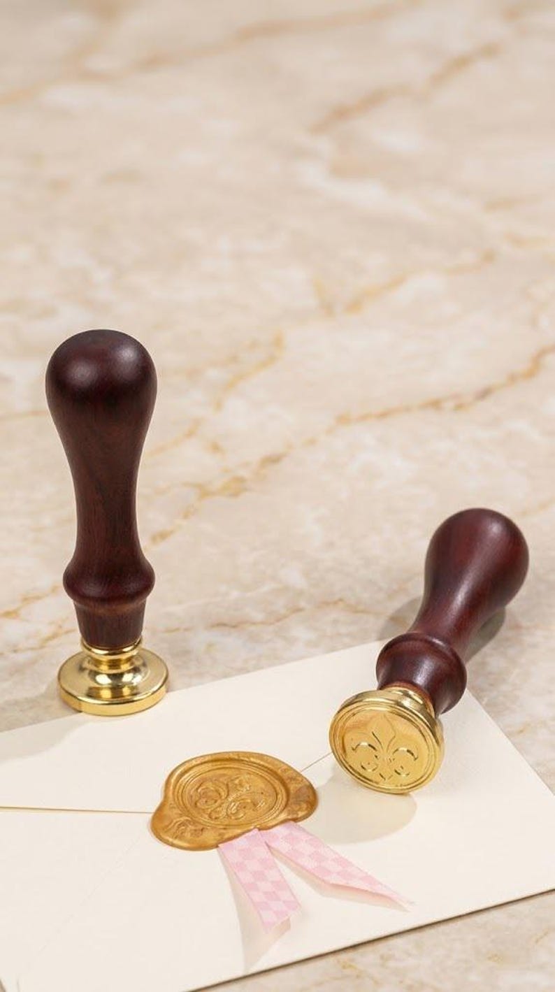Op de afbeelding: Twee houten lakstempels met goudkleurige bases en donkerbruine handvatten. E&eacute;n stempel staat rechtop, de andere ligt op een cr&egrave;mekleurige envelop met een gouden lakzegel en roze lint. De zegel heeft een bloemenmotief.