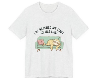 May include: A light grey short-sleeve t-shirt featuring a graphic of a sloth sleeping on a light green sofa next to a pink pillow. Above the sofa, the text reads: I'VE REACHED MY LIMIT (IT WAS LOW). The design is centred on the chest area.