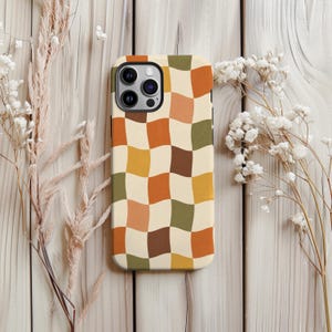 Peut inclure: Une coque de smartphone avec un motif à damier ondulé répétitif dans des tons crème, jaune moutarde, orange brûlé, vert olive et brun foncé. La coque présente une découpe pour un système de caméra à trois objectifs en haut à gauche.