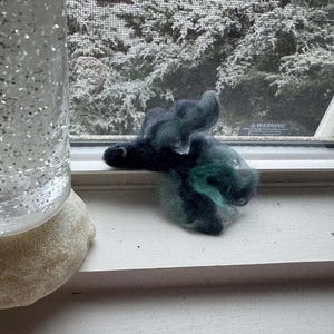 May include: A handmade felted sculpture in shades of black and teal, resembling a stylized animal head, rests on a white windowsill. A glitter-filled glass snow globe is partially visible to the left. The background shows a snowy outdoor scene.