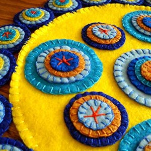 K&ouml;nnte beinhalten: Ein gelber Filzkreis mit mehreren konzentrischen Kreisen aus blauem, orangefarbenem und gelbem Filz, die darauf gen&auml;ht sind. Die Kreise sind kreisf&ouml;rmig um den gelben Filzkreis angeordnet.