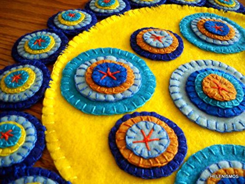 K&ouml;nnte beinhalten: Ein gelber Filzkreis mit mehreren konzentrischen Kreisen aus blauem, orangefarbenem und gelbem Filz, die darauf gen&auml;ht sind. Die Kreise sind kreisf&ouml;rmig um den gelben Filzkreis angeordnet.