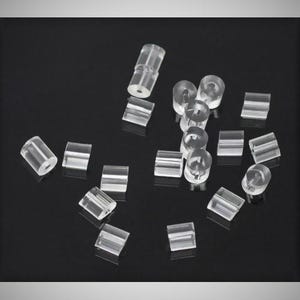 May include: A collection of small, clear, cylindrical plastic beads scattered across the frame. Each bead features a smooth, transparent surface and a central hole running through the length of the cylinder, creating a uniform, translucent appearance.