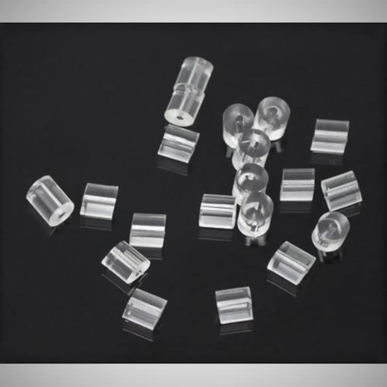 May include: A collection of small, clear, cylindrical plastic beads scattered across the frame. Each bead features a smooth, transparent surface and a central hole running through the length of the cylinder, creating a uniform, translucent appearance.