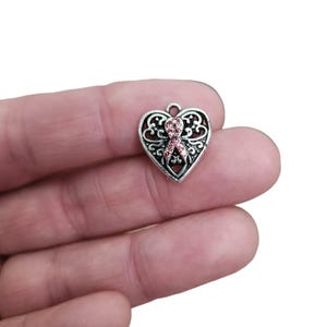 May include: A silver-colored heart-shaped charm with an open filigree design. A pink ribbon made of small crystals is centered on the charm. The charm has a small loop at the top for attaching to a necklace or bracelet. The charm is held in a hand.