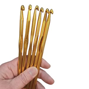 May include: A set of six gold crochet hooks, all the same size. The hooks are held in a hand, and the text "6.0mm" is visible on one of the hooks.