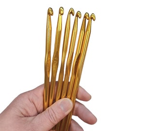 May include: A set of six gold crochet hooks, all the same size. The hooks are held in a hand, and the text "6.0mm" is visible on one of the hooks.