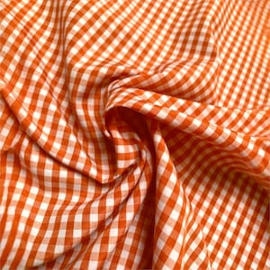 Puede incluir: Primer plano de tela de cuadros naranja y blanca. La tela está doblada y drapeada, mostrando el patrón de cuadrados espaciados uniformemente. El material parece ser algodón o un textil tejido similar, adecuado para varios proyectos de costura.
