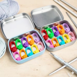 May include: Two open metal tins filled with colorful stitch markers. The markers are in various colors, including red, green, purple, orange, and yellow. The tins are next to a ball of gray yarn and knitting needles, suggesting a knitting or crochet project.