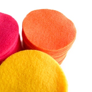 May include: Stacks of colorful felt circles in shades of pink, orange, and yellow. The felt circles are neatly arranged, with each stack showing the texture and vibrant colors of the material. The image is set against a clean white background.
