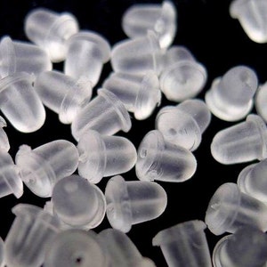 May include: A close-up of a pile of clear plastic earring backs. The backs are small and round, with a slightly pointed top. They are designed to be placed on the back of an earring to keep it in place.