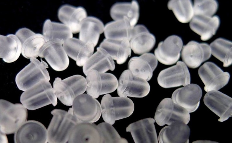 May include: A close-up of a pile of clear plastic earring backs. The backs are small and round, with a slightly pointed top. They are designed to be placed on the back of an earring to keep it in place.