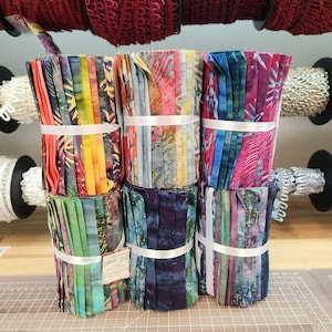 May include: Bundles of colorful fabric strips, tied with white ribbon. The fabric features a variety of patterns and colors, including orange, yellow, green, blue, and purple. Rolls of lace and trim are visible in the background.