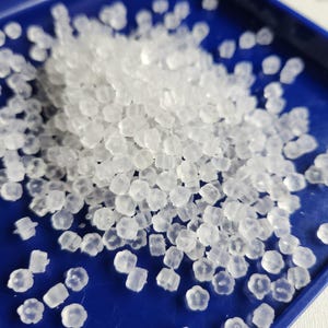 May include: A collection of small, translucent, flower-shaped earring backs made of soft silicone. These tiny, clear, petal-patterned components are scattered across a smooth, deep blue surface, highlighting their distinct floral shape and texture.