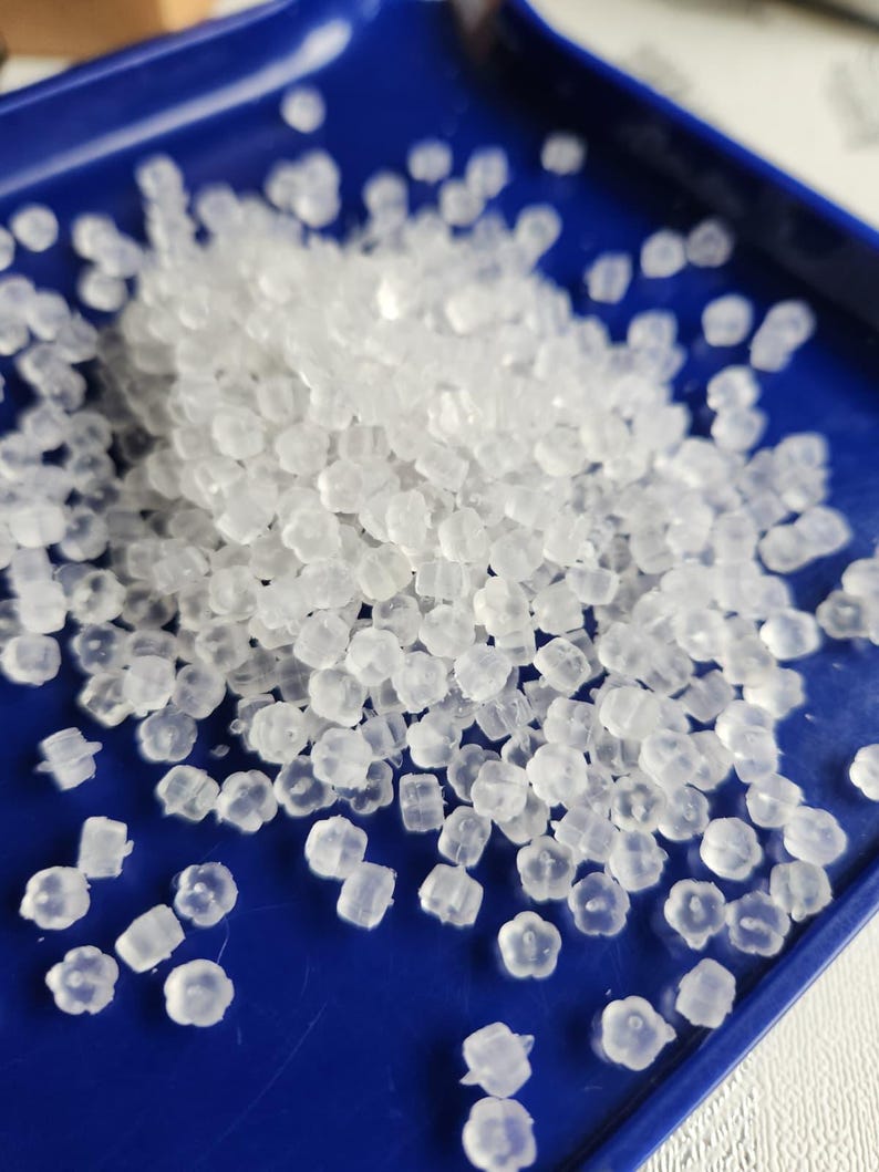 May include: A collection of small, translucent, flower-shaped earring backs made of soft silicone. These tiny, clear, petal-patterned components are scattered across a smooth, deep blue surface, highlighting their distinct floral shape and texture.
