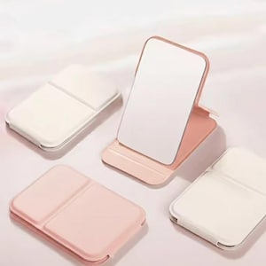 Peut inclure: Quatre petits miroirs cosmétiques pliables, de couleurs rose et blanc. L'un est ouvert, montrant la surface réfléchissante, tandis que les autres sont fermés. Design minimaliste, idéal pour les voyages ou le quotidien.