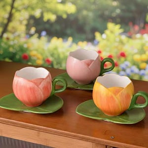 Op de afbeelding: Drie keramische theekopjes in tulpenvorm in roze en gele tinten, elk met een groen handvat en rustend op een groene, bladvormige schotel. De kopjes staan op een houten oppervlak met een vage bloemenachtergrond.