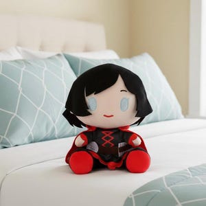 Peut inclure: Une poupée en peluche avec une coupe au carré noire, des yeux bleu clair et une petite bouche rouge. La poupée porte une tenue noire et rouge avec une cape rouge et des chaussures rouges. La poupée est assise sur un lit blanc avec des oreillers bleu clair.