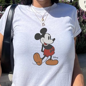 Puede incluir: Camiseta blanca con un gr&aacute;fico de Mickey Mouse en negro, rojo y marr&oacute;n. El logotipo de Disney est&aacute; impreso debajo del personaje. La camiseta tiene mangas cortas y cuello redondo.