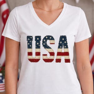 Könnte beinhalten: Ein weißes kurzärmeliges T-Shirt mit V-Ausschnitt und dem Schriftzug USA auf der Brust. Die Buchstaben sind mit einem abgenutzten amerikanischen Flaggenmuster gefüllt, das rote und weiße horizontale Streifen sowie ein blaues Feld mit weißen Sternen zeigt.