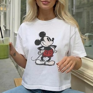 Puede incluir: Una camiseta blanca de manga corta con una ilustraci&oacute;n gr&aacute;fica centrada de Mickey Mouse. El personaje est&aacute; representado en un estilo cl&aacute;sico con orejas negras, cuerpo negro y pantalones cortos rojos. La camiseta tiene cuello redondo.