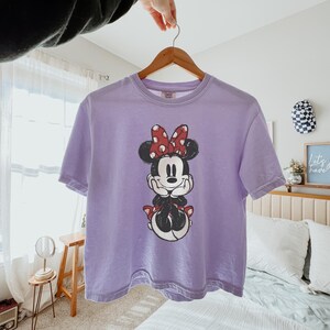 K&ouml;nnte beinhalten: Ein hellviolettes Kurzarm-T-Shirt mit einem Grafikdruck von Minnie Maus auf der Vorderseite. Die Figur ist mit einer rot-wei&szlig; gepunkteten Schleife, schwarzen Ohren sowie einem schwarz-wei&szlig;en K&ouml;rper dargestellt und st&uuml;tzt das Kinn auf die H&auml;nde.