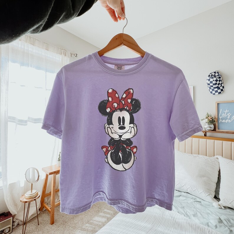 K&ouml;nnte beinhalten: Ein hellviolettes Kurzarm-T-Shirt mit einem Grafikdruck von Minnie Maus auf der Vorderseite. Die Figur ist mit einer rot-wei&szlig; gepunkteten Schleife, schwarzen Ohren sowie einem schwarz-wei&szlig;en K&ouml;rper dargestellt und st&uuml;tzt das Kinn auf die H&auml;nde.