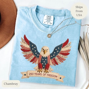 May include: Light blue t-shirt with an embroidered eagle design. The eagle's wings feature red, white, and blue fabric patches. The shirt has the text "250 YEARS OF FREEDOM" on a banner below the eagle. The shirt is labeled "Chambray".