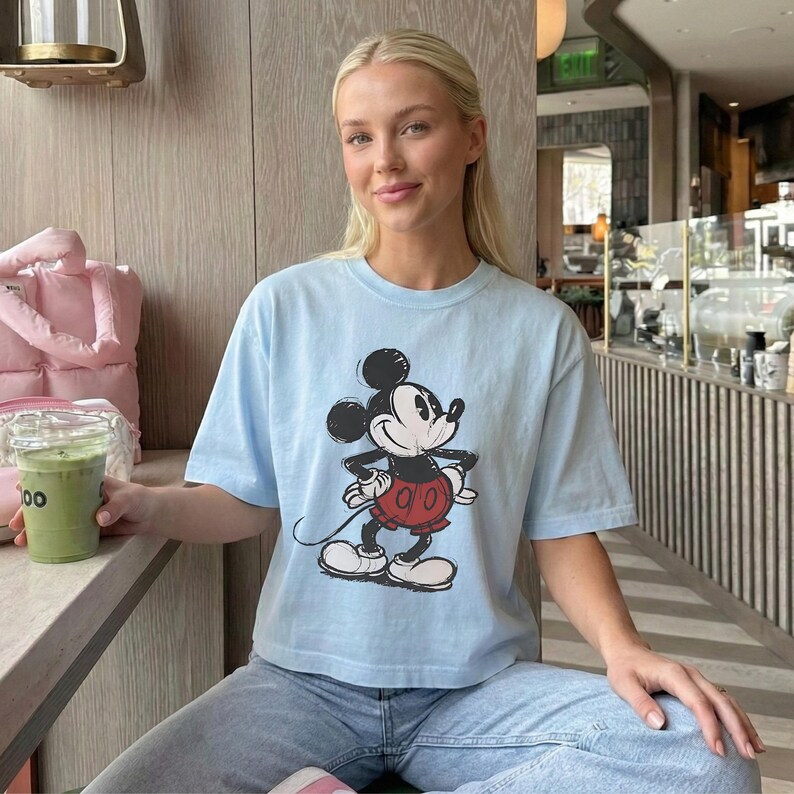 Puede incluir: Una camiseta de manga corta de color azul claro con un estampado gr&aacute;fico de Mickey Mouse en la parte delantera. El personaje aparece en un estilo cl&aacute;sico con orejas negras, cuerpo negro, pantalones cortos rojos y guantes y zapatos blancos.