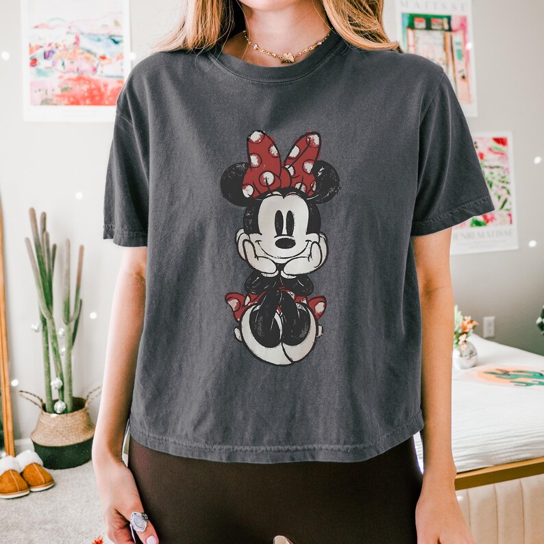 K&ouml;nnte beinhalten: Ein anthrazitfarbenes Kurzarm-T-Shirt mit einem verwaschenen Grafikdruck von Minnie Maus. Die Figur ist mit ihrer charakteristischen rot-wei&szlig;en gepunkteten Schleife, schwarzen Ohren und wei&szlig;en Handschuhen dargestellt, das Kinn auf die H&auml;nde gest&uuml;tzt.