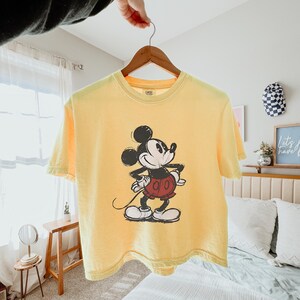 Puede incluir: Una camiseta de manga corta de color amarillo p&aacute;lido con un gr&aacute;fico de estilo vintage de Mickey Mouse en el centro. El personaje aparece en negro, blanco y rojo, de pie con las manos en la cintura. La camiseta cuelga de una percha de madera.