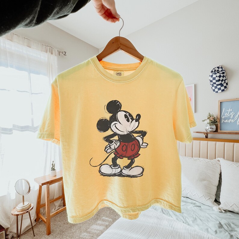 Puede incluir: Una camiseta de manga corta de color amarillo p&aacute;lido con un gr&aacute;fico de estilo vintage de Mickey Mouse en el centro. El personaje aparece en negro, blanco y rojo, de pie con las manos en la cintura. La camiseta cuelga de una percha de madera.
