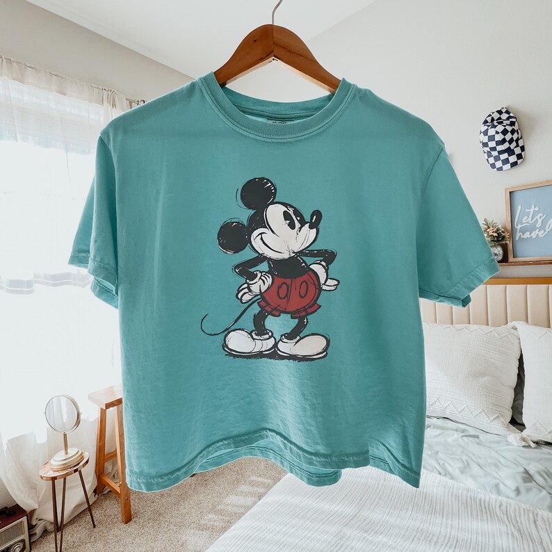 Puede incluir: Una camiseta de manga corta color verde azulado con un estampado gr&aacute;fico desgastado de Mickey Mouse en el centro. El personaje aparece en negro, blanco y rojo, de pie con las manos en la cintura. La camiseta cuelga de una percha de madera.