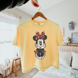 K&ouml;nnte beinhalten: Ein hellgelbes Kurzarm-T-Shirt mit einer mittigen Grafik von Minnie Maus, die ihre H&auml;nde unter das Kinn st&uuml;tzt. Die Figur ist im schwarz-wei&szlig;en Skizzenstil gehalten und tr&auml;gt eine rote Schleife mit wei&szlig;en Punkten auf dem Kopf.