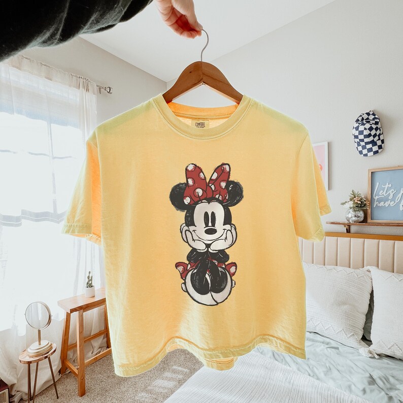 K&ouml;nnte beinhalten: Ein hellgelbes Kurzarm-T-Shirt mit einer mittigen Grafik von Minnie Maus, die ihre H&auml;nde unter das Kinn st&uuml;tzt. Die Figur ist im schwarz-wei&szlig;en Skizzenstil gehalten und tr&auml;gt eine rote Schleife mit wei&szlig;en Punkten auf dem Kopf.