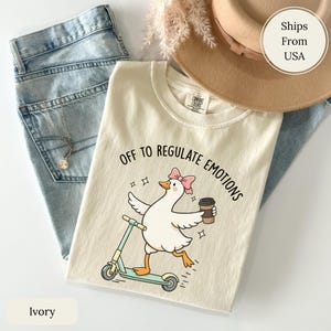 Peut inclure: T-shirt ivoire avec l'inscription "Off to Regulate Emotions" et un dessin de canard sur un scooter, tenant une tasse de café. Le t-shirt est présenté avec un jean bleu et un chapeau beige.