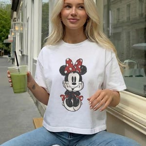 K&ouml;nnte beinhalten: Ein wei&szlig;es Kurzarm-T-Shirt mit einer mittigen Grafik von Minnie Maus, die eine rot-wei&szlig; gepunktete Schleife und schwarze Ohren tr&auml;gt. Der Druck ist im abgenutzten Vintage-Stil gehalten und zeigt die Figur mit H&auml;nden unter dem Kinn.
