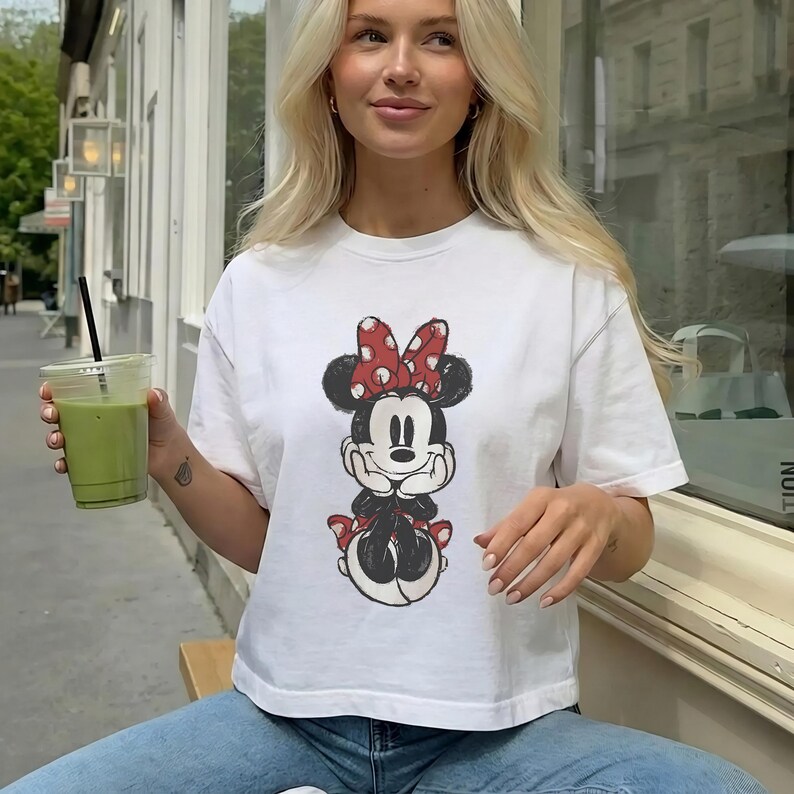 K&ouml;nnte beinhalten: Ein wei&szlig;es Kurzarm-T-Shirt mit einer mittigen Grafik von Minnie Maus, die eine rot-wei&szlig; gepunktete Schleife und schwarze Ohren tr&auml;gt. Der Druck ist im abgenutzten Vintage-Stil gehalten und zeigt die Figur mit H&auml;nden unter dem Kinn.