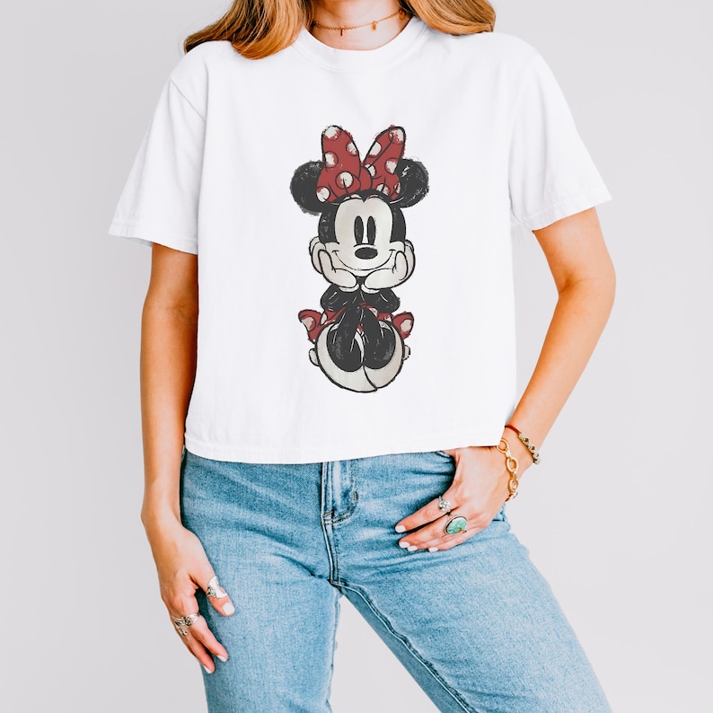 K&ouml;nnte beinhalten: Ein wei&szlig;es Kurzarm-T-Shirt mit einer mittigen Grafik von Minnie Maus in Used-Optik. Die Figur ist mit einer rot-wei&szlig;en gepunkteten Schleife, schwarzen Ohren sowie einem schwarz-wei&szlig;en K&ouml;rper dargestellt und st&uuml;tzt ihr Kinn auf ihre H&auml;nde.