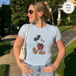 Puede incluir: Camiseta azul claro con un dibujo de Mickey Mouse en negro, rojo y marr&oacute;n. El personaje de dibujos animados est&aacute; representado de pie. La camiseta se lleva con unos vaqueros azules. La imagen est&aacute; tomada al aire libre.