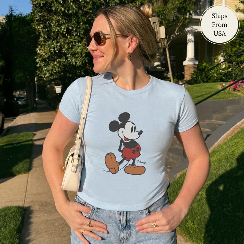 Puede incluir: Camiseta azul claro con un dibujo de Mickey Mouse en negro, rojo y marr&oacute;n. El personaje de dibujos animados est&aacute; representado de pie. La camiseta se lleva con unos vaqueros azules. La imagen est&aacute; tomada al aire libre.