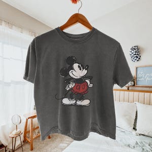 Puede incluir: Una camiseta de manga corta en color gris carb&oacute;n con un gr&aacute;fico de estilo vintage de Mickey Mouse. El personaje aparece en blanco y negro con pantalones cortos rojos, de pie con las manos en la cintura. El dise&ntilde;o tiene un aspecto desgastado.