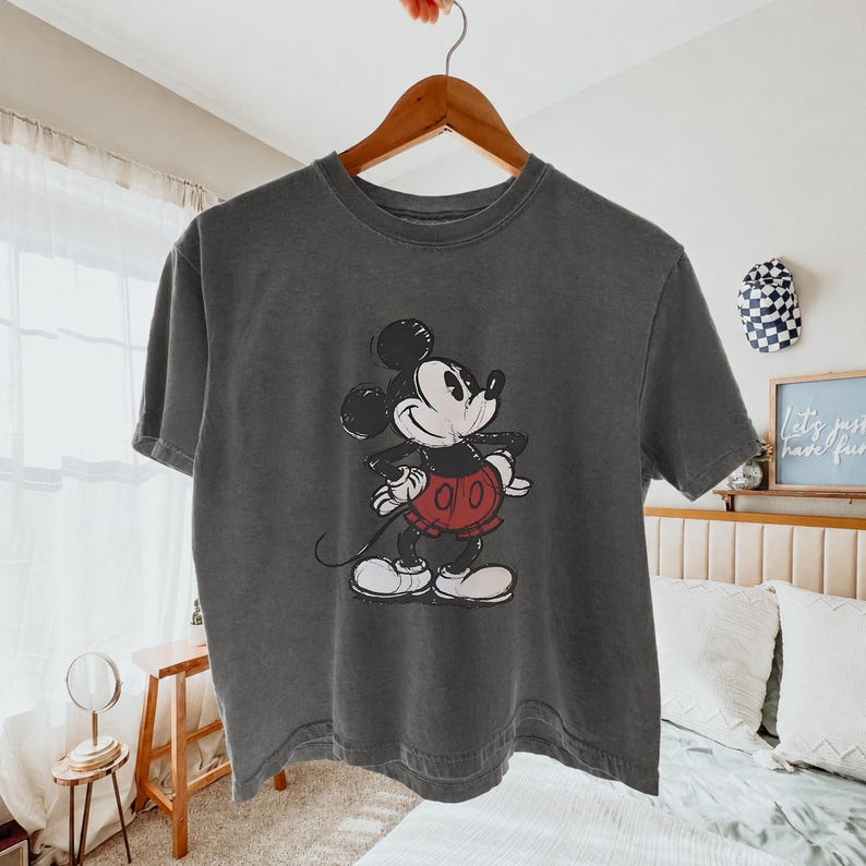 Puede incluir: Una camiseta de manga corta en color gris carb&oacute;n con un gr&aacute;fico de estilo vintage de Mickey Mouse. El personaje aparece en blanco y negro con pantalones cortos rojos, de pie con las manos en la cintura. El dise&ntilde;o tiene un aspecto desgastado.