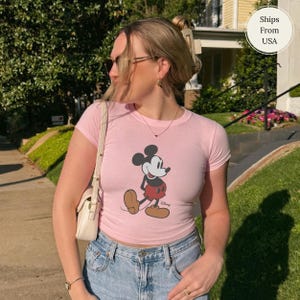 Puede incluir: Camiseta rosa claro con un dibujo de Mickey Mouse caminando. La camiseta tiene mangas cortas y cuello redondo. La persona lleva vaqueros azules y un bolso blanco.