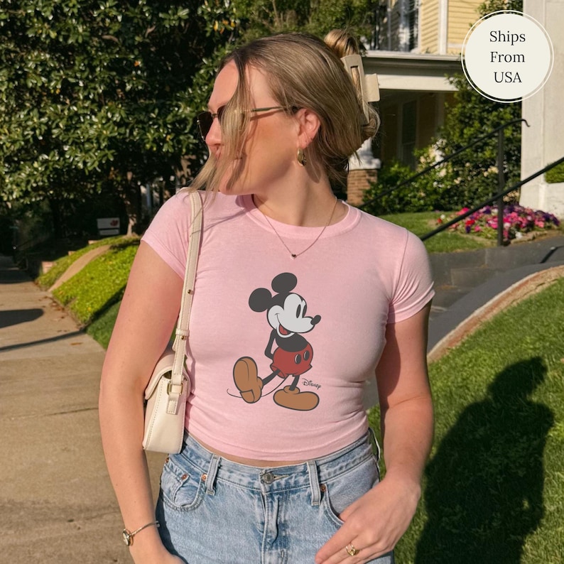 Puede incluir: Camiseta rosa claro con un dibujo de Mickey Mouse caminando. La camiseta tiene mangas cortas y cuello redondo. La persona lleva vaqueros azules y un bolso blanco.