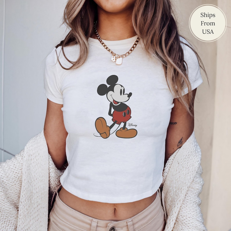 Puede incluir: Top corto blanco con un gr&aacute;fico de Mickey Mouse en negro, rojo y marr&oacute;n. El personaje de Disney se representa caminando. La camiseta tiene mangas cortas y cuello redondo.