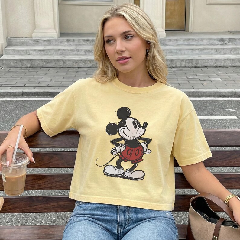 Puede incluir: Una camiseta de manga corta de color amarillo p&aacute;lido con un gr&aacute;fico de estilo vintage de Mickey Mouse en negro, blanco y rojo. El personaje aparece en una pose cl&aacute;sica con las manos en la cintura sobre el tejido amarillo claro liso.