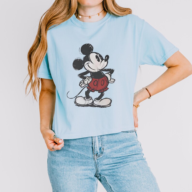 Puede incluir: Una camiseta de manga corta de color azul claro con un gr&aacute;fico de estilo boceto en blanco y negro de Mickey Mouse con pantalones cortos rojos. El personaje aparece en una pose cl&aacute;sica con las manos en la cintura y textura de tinta.