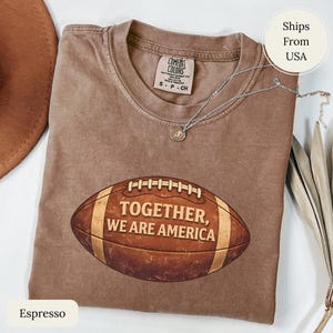 May include: Espresso-coloured t-shirt featuring a football graphic with the text "TOGETHER, WE ARE AMERICA." The shirt has a crew neck and short sleeves. A silver necklace with a small pendant is draped across the shirt. The shirt is a size small.