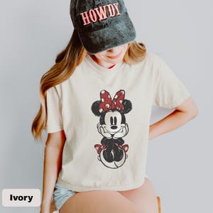 K&ouml;nnte beinhalten: Ein elfenbeinfarbenes Kurzarm-T-Shirt mit einer Grafik einer Zeichentrickmaus mit roter, gepunkteter Schleife, kombiniert mit einer verwaschenen schwarzen Baseballkappe, auf deren Vorderseite das Wort HOWDY in roten Buchstaben gestickt ist.